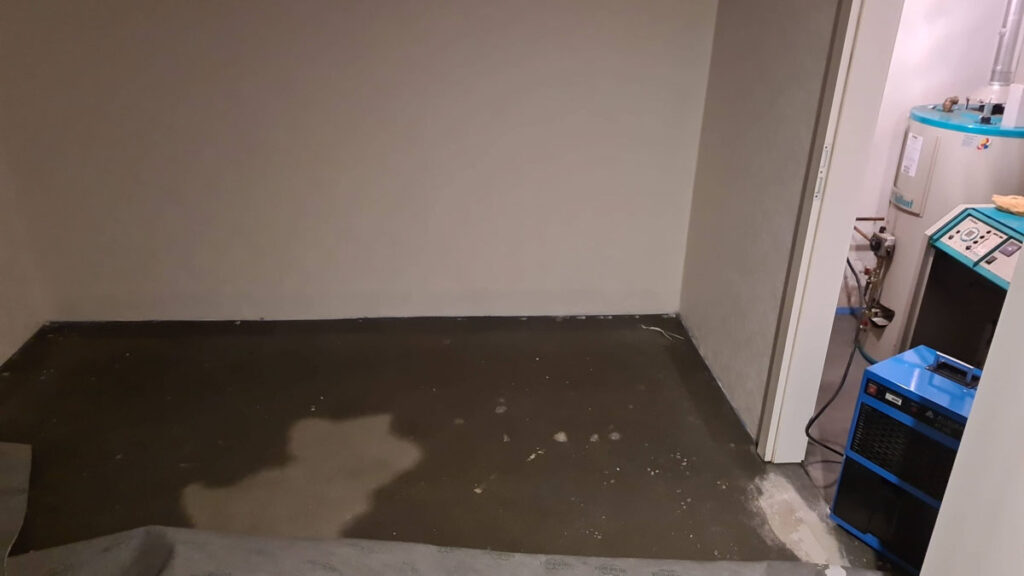 Ein kellerraum mit nassem betonboden, sichtbaren wasserschäden und einem blauen luftentfeuchter neben einem warmwasserbereiter in einem angrenzenden wirtschaftsraum. die wände sind blass und schlicht.