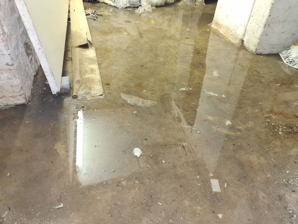 Ein kellerraum mit einem betonboden, der mit stehendem wasser bedeckt ist und anzeichen einer Überschwemmung aufweist. baumaterial und schutt liegen verstreut herum, und die wände scheinen unfertig zu sein.