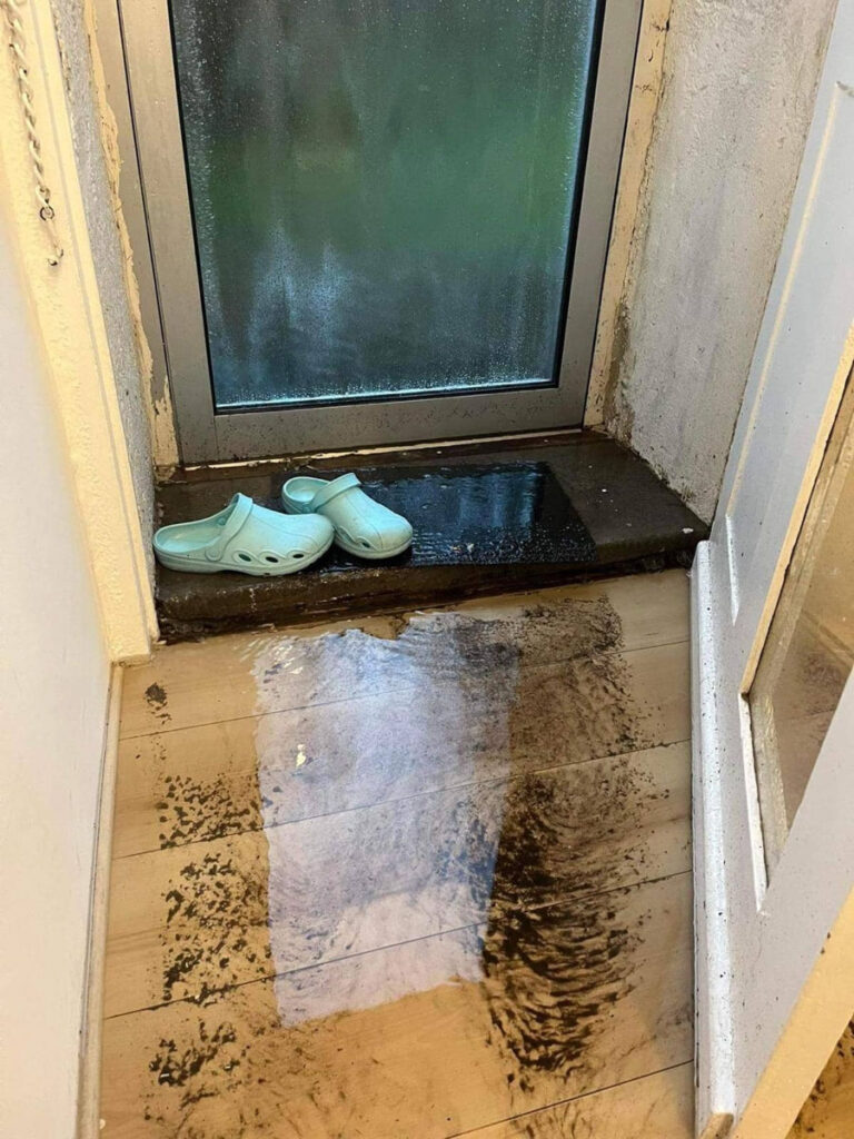 Ein kleiner eingang mit schlammigem wasser, das einen teil des holzbodens bedeckt, nasse fußspuren, die von einer glastür wegführen, und ein paar hellblaue clogs, die auf der schwelle stehen.