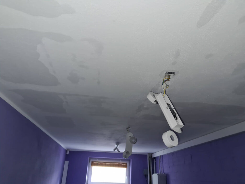 Ein raum mit violetten wänden und einem fenster am anderen ende; die decke weist wasserschäden auf, und eine kaputte deckenleuchte mit freiliegenden kabeln hängt herunter.