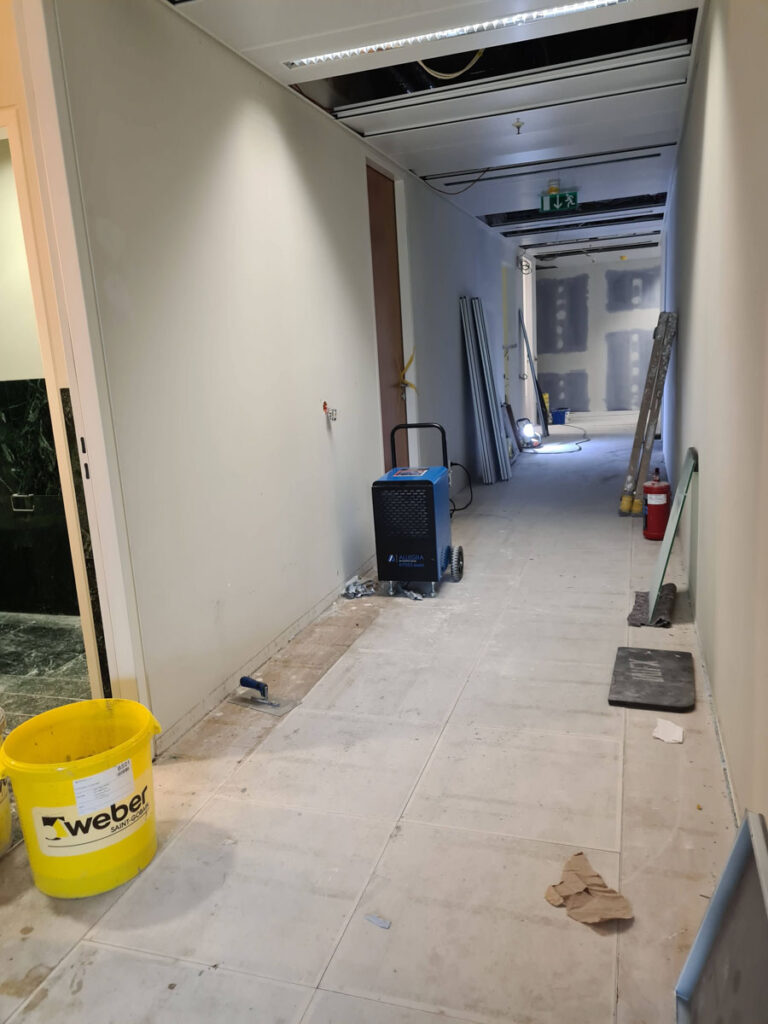 Ein im bau befindlicher flur mit freiliegenden deckenplatten, baumaterialien, einem gelben eimer, einem tragbaren luftkissengerät, leitern und unfertigen wänden und böden.