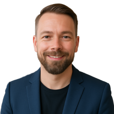 Ein lächelnder Mann mit kurzen hellbraunen Haaren und einem Bart, der einen marineblauen Blazer über einem schwarzen Hemd trägt, posiert vor einem weißen Hintergrund.