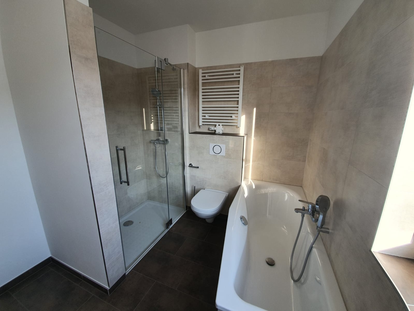 Modernes Badezimmer mit Glasduschkabine, wandmontierter Toilette, weißer Badewanne mit Duschkopf, beheiztem Handtuchhalter und beigefarbenen Fliesen an Wand und Boden. Durch ein Fenster neben der Badewanne fällt Sonnenlicht ein.