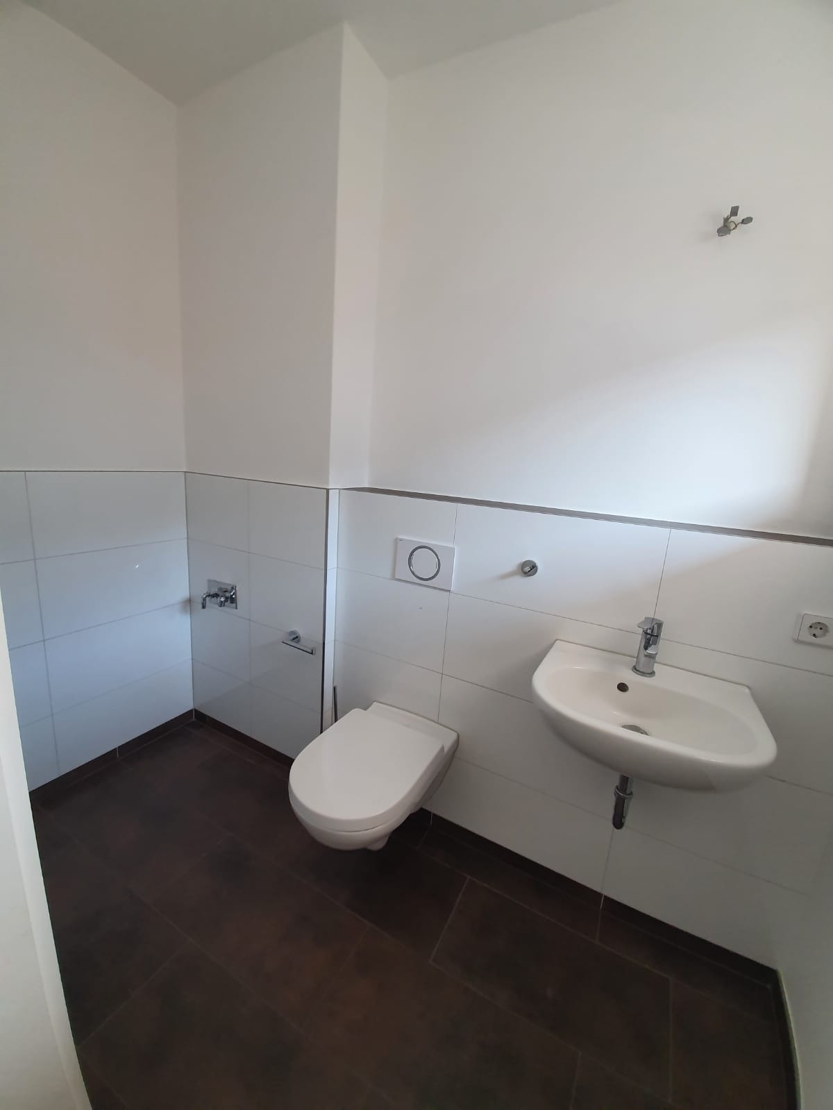 Ein minimalistisches Badezimmer mit weiß gefliesten Wänden, einer wandmontierten Toilette, einem kleinen wandmontierten Waschbecken und einem braunen Fliesenboden. Es gibt einen Toilettenpapierhalter und einen Wasserhahn, aber keinen sichtbaren Spiegel oder andere Accessoires.