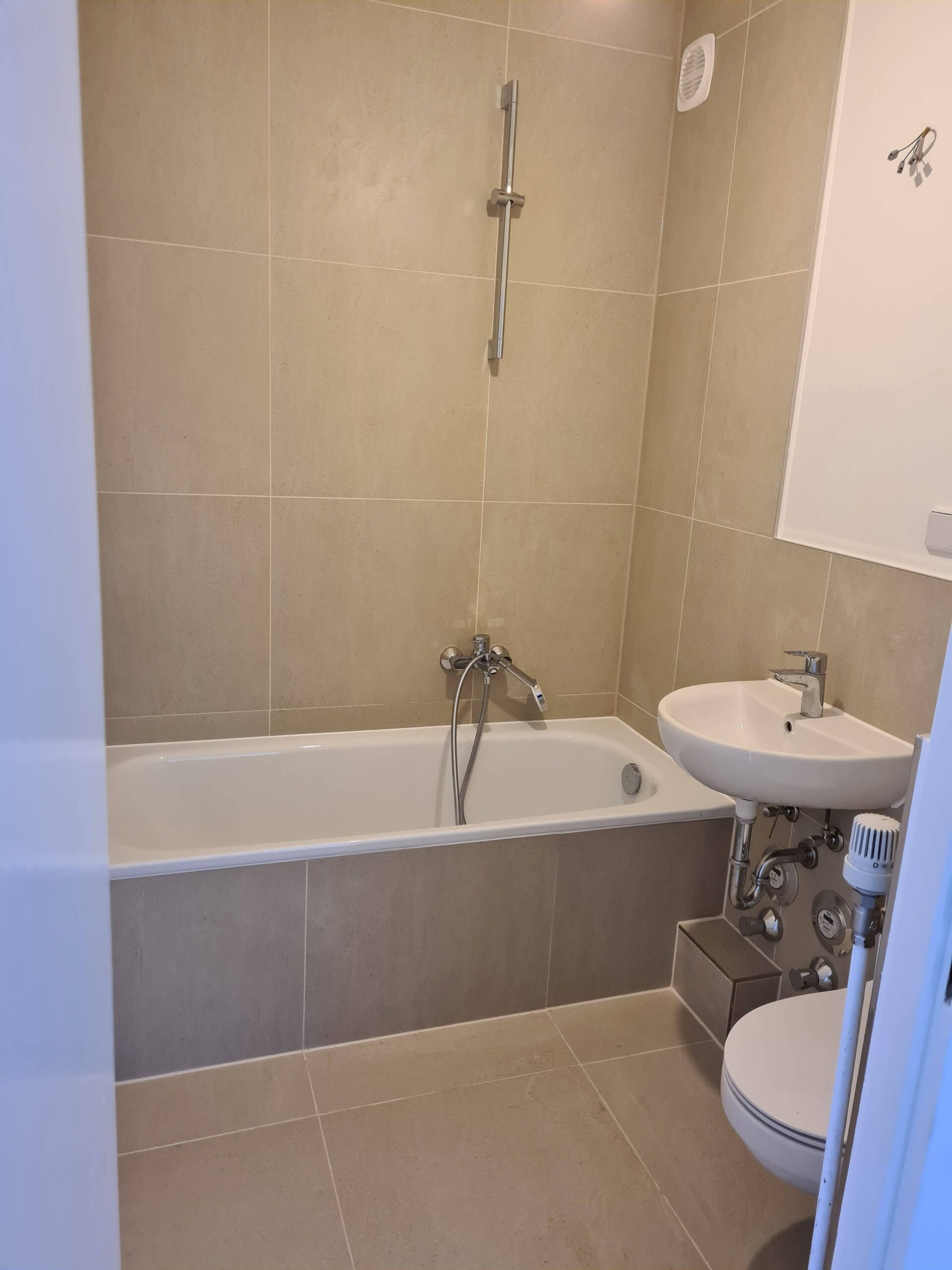 Ein beige gefliestes Badezimmer mit einer weißen Badewanne und einer an der Wand befestigten Dusche, einem kleinen Waschbecken mit freiliegenden Rohrleitungen und einer Teilansicht einer Toilette in der unteren rechten Ecke.