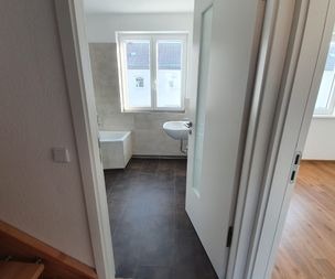 Ein Badezimmer mit einer weißen, teilweise geöffneten Tür, die den Blick auf eine Badewanne, ein an der Wand befestigtes Waschbecken und zwei große Fenster freigibt. Der Boden ist mit dunklen Fliesen ausgelegt, und der Raum ist von natürlichem Licht durchflutet. Daneben befindet sich ein Raum mit Holzfußboden.