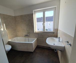 Ein helles Badezimmer mit einem großen Fenster, einer weißen Badewanne in der Ecke, einem an der Wand befestigten weißen Waschbecken und einer Toilette. Die Wände sind teilweise beige gefliest, der Boden ist dunkelbraun.