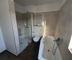 Modernes, beige gefliestes Bad mit Glasdusche, Wandtoilette unter einem Handtuchheizkörper und weißer Badewanne mit verchromtem Wasserhahn neben einem Fenster.