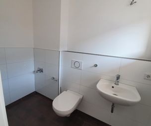 Ein minimalistisches Badezimmer mit weiß gefliesten Wänden, einer wandmontierten Toilette, einem kleinen Wandwaschbecken mit Wasserhahn und dunklen Bodenfliesen. Der Raum wirkt sauber und aufgeräumt.