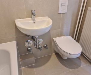 Ein kleines Wandwaschbecken mit freiliegenden Rohrleitungen ist neben einer Wandtoilette in einem modernen Badezimmer mit beigen Fliesen installiert. Der Rand einer Badewanne und ein weißer Heizkörper sind ebenfalls sichtbar.