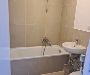 Ein kleines Badezimmer mit beige gefliesten Wänden, einer weißen Badewanne mit einer an der Wand befestigten Duschbrause und einem kompakten weißen Waschbecken mit freiliegenden Rohrleitungen darunter.