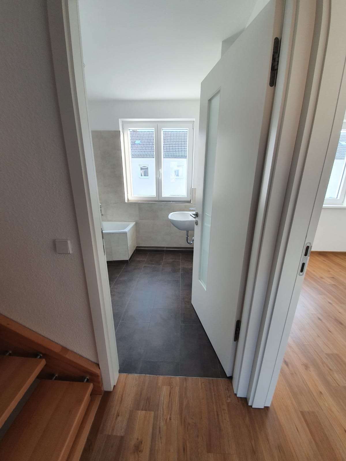 Blick in ein helles Badezimmer mit einem Fenster, einem an der Wand befestigten Waschbecken und einer Badewanne. Die Tür ist offen und gibt den Blick auf dunkle Bodenfliesen im Bad und Holzböden außerhalb des Zimmers frei. Auf der linken Seite sind Treppenstufen zu sehen.