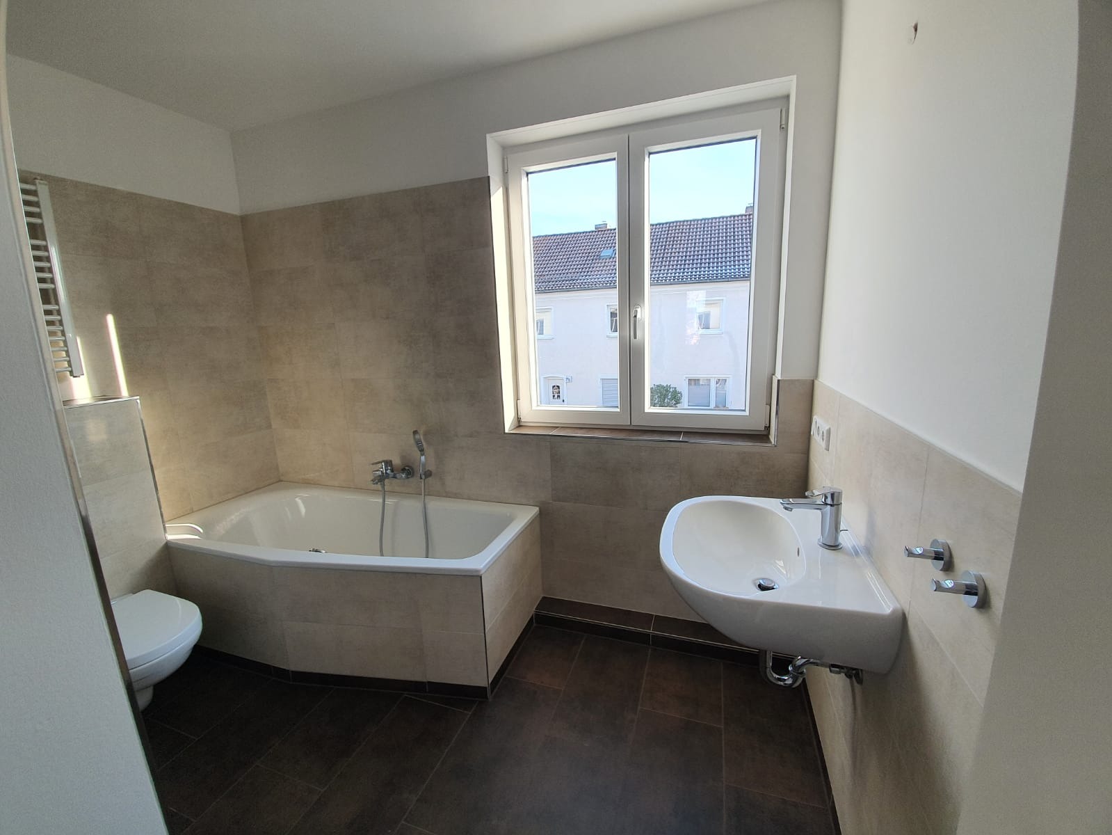 Ein modernes Badezimmer mit weißer Badewanne, wandmontiertem Waschbecken und Toilette. Die Wände sind beigefarben gefliest, und ein großes Fenster lässt viel Tageslicht herein. Der Boden ist dunkelbraun.