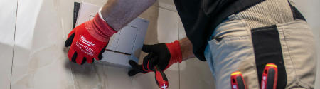 Eine Person mit roten Handschuhen verlegt mit einem Schraubenzieher eine Fliese an einer Wand, wobei sie das Werkzeug sichtbar in der Hosentasche verstaut.
